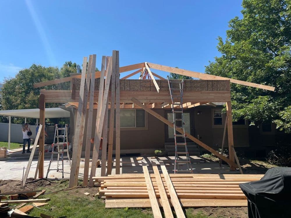 Home under construction with wooden framework, ladders, and stacked timber in a sunny backyard.
