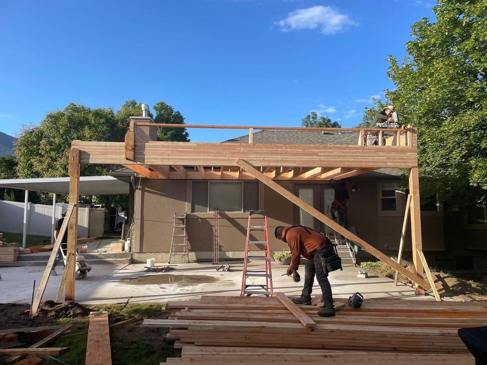 Construction workers building a wooden deck on a residential home, with tools and materials visible.